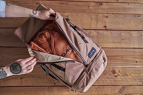 Person packing a Dakine bag with an orange hoodie on a wooden surface.